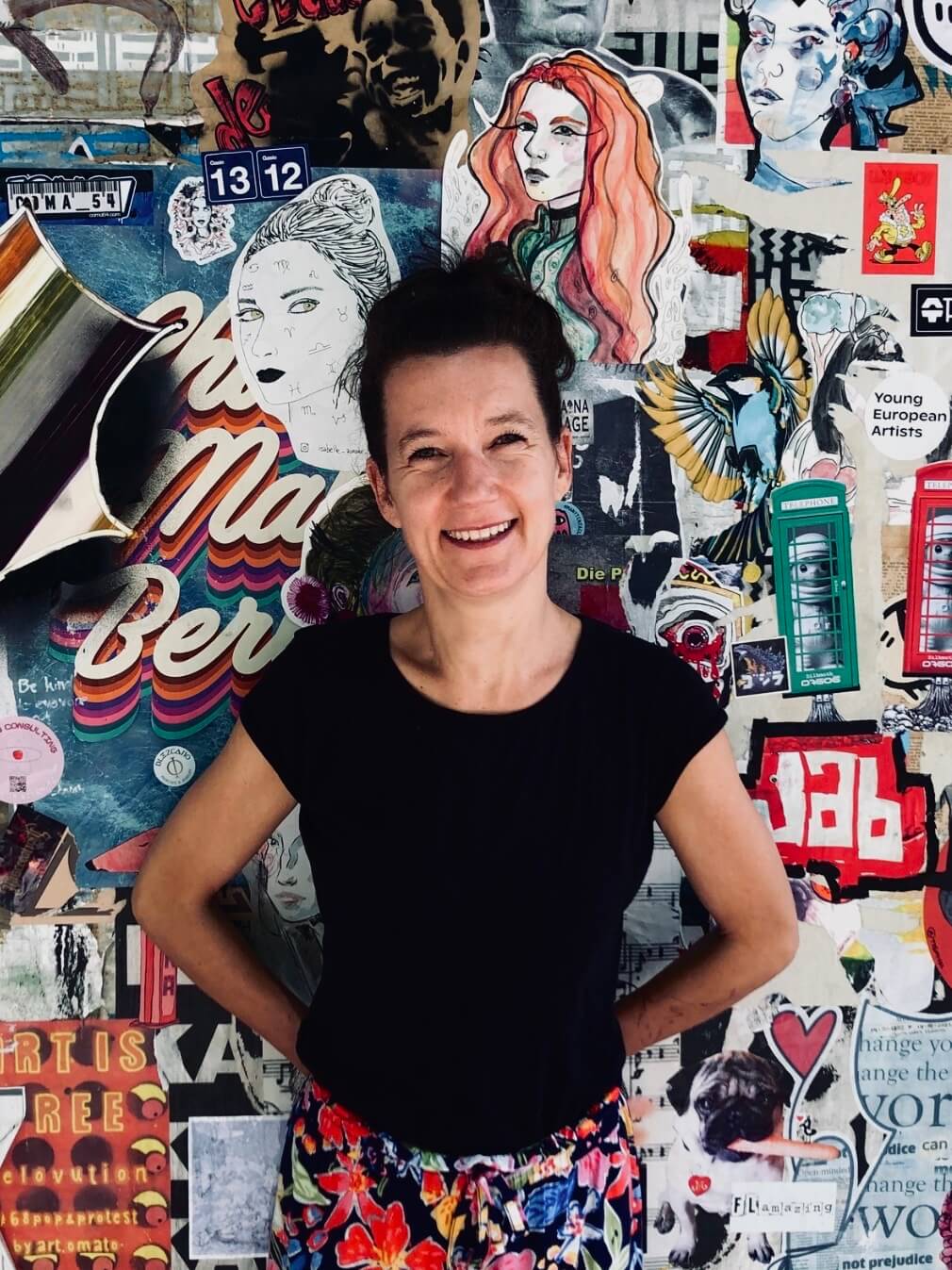 Susanne_Ennis_Virtuelle_Assitenz_Podcast Frau mit schwarzem Shirt und bunter Hose steht lächelnd vor einer kreativen Wand voller Kunst und Street-Art-Collagen.